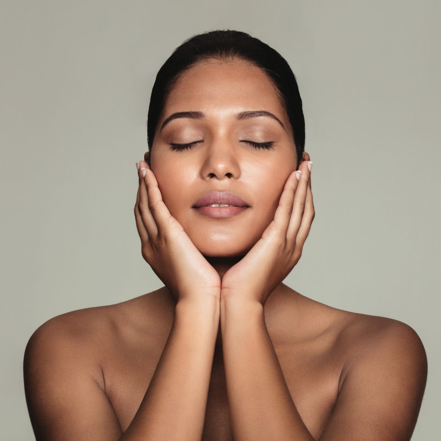 Portrait of young natural beauty with beautiful skin. Female touching her fresh clean and clear face against grey background.