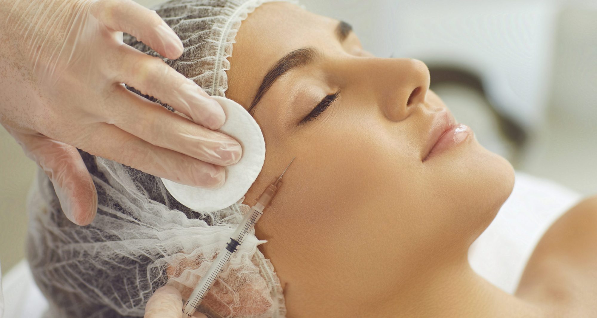 Cosmetologist wiping skin with cotton pads during facial beauty botox injection for young womans cheek in beauty salon, side view. Facial treatment and beauty injection concept